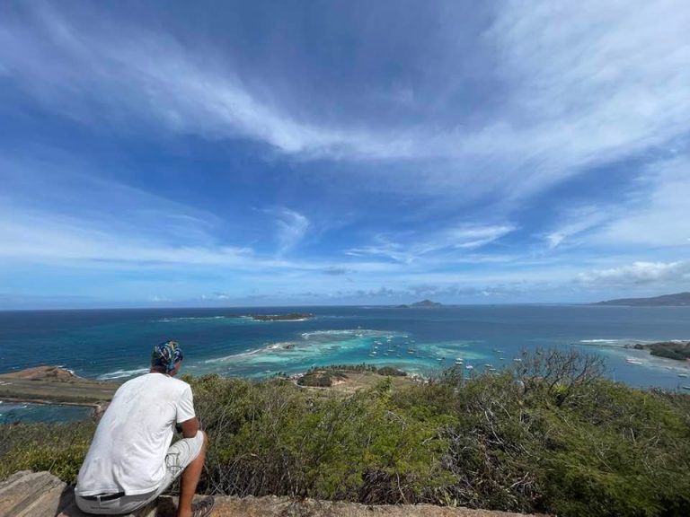 A man sitting on a headland looking out over turquoise water and small islands.