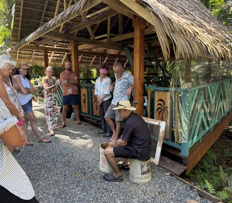 A group of visitors gathered under a thatched shelter on a gravel path, with a man seated on a bucket.