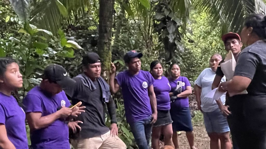 A group of people in purple shirts listening to a speaker in the forest.