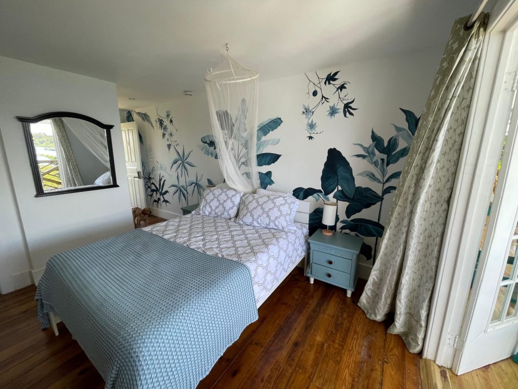 A bedroom with a mosquito net above the bed, a painted plant mural and doors to the balcony