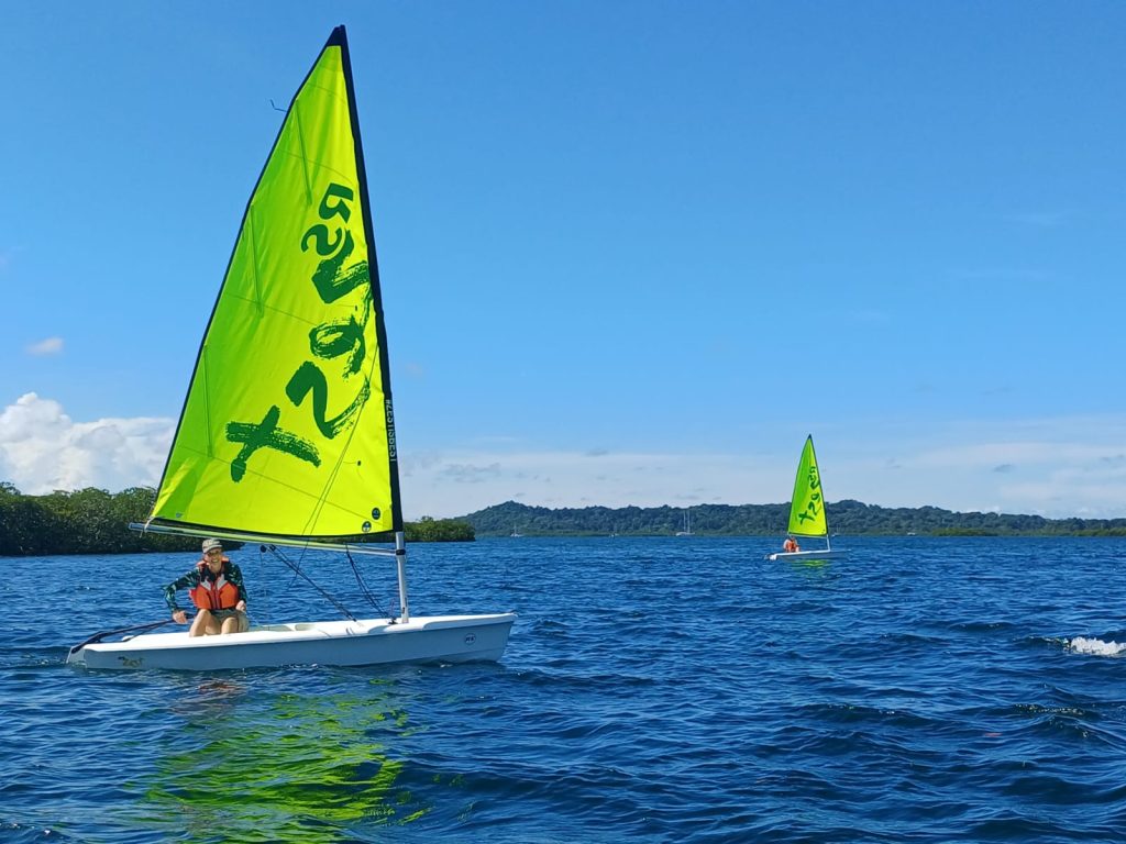 Two Zest sailing dinghies with bright lime green sails on the blue water of Bahía Honda. A smiling sailor in an orange life jacket sails the nearer boat, with mangroves and forested hills behind.